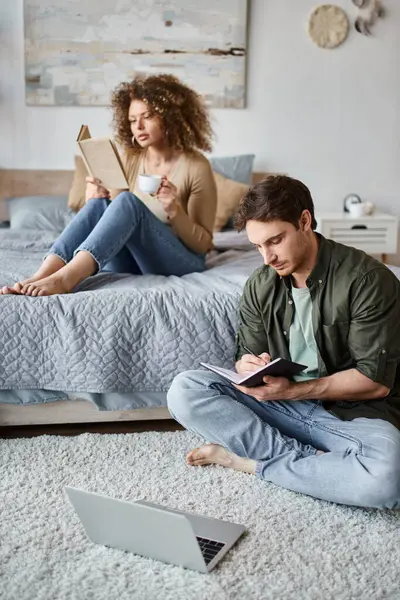 Couple spending time at home. Woman reading book on bed and man is taking notes sitting on the floor — Stock Photo