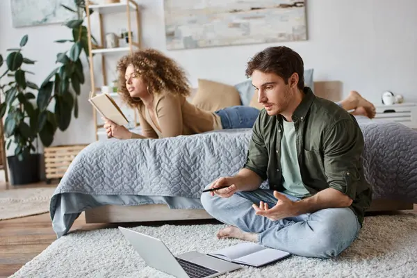 Couple working remotely. Woman reading book on bed and man is online chatting sitting on the floor — Stock Photo