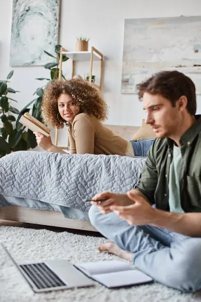 Couple working remotely. Woman reading book on bed and looking at man online chatting on the floor — Stock Photo