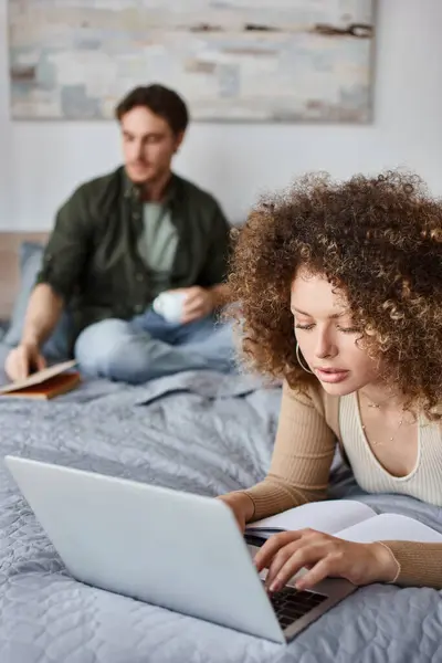 Couple à la maison. Femme utilise un ordinateur portable tandis que l'homme est assis avec livre et tasse de café sur le lit — Photo de stock