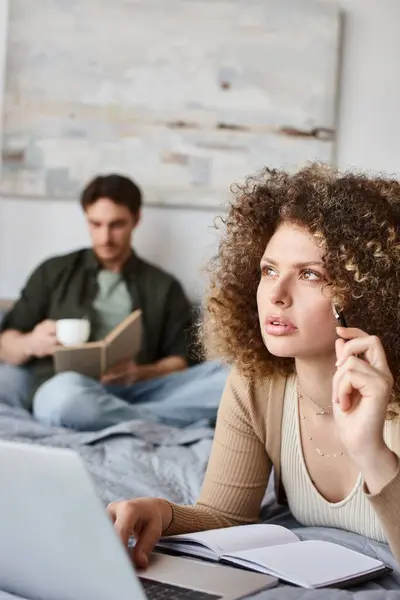 Couple working at home. Concerned woman is working with laptop while man is reading book on bed — Stock Photo