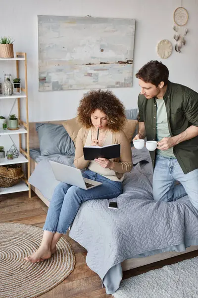 Curly young woman and brunette man sitting cozily in bed, man holding coffee cups - foto de stock