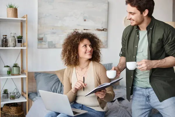 Curly young woman and brunette man sitting cozily in bed, man offering coffee to his partner — Stock Photo