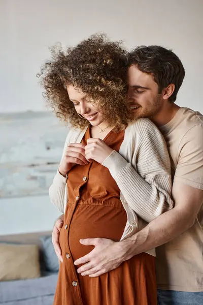 Прекрасный счастливый мужчина обнимает беременную жену сзади, стоя дома и ожидая ребенка. — стоковое фото