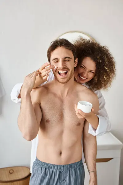 Happy couple in love in bathroom. Woman applying cream to her man in bathroom and laughing — Stock Photo