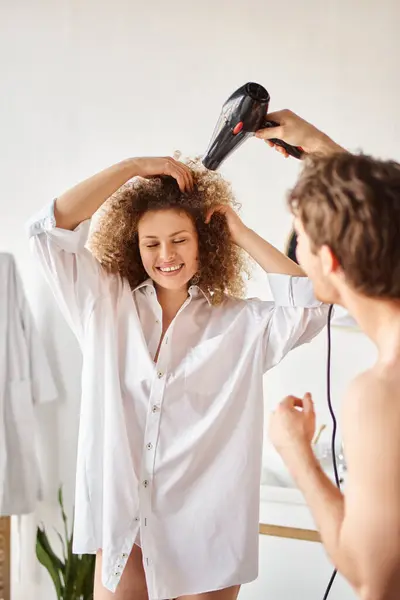 Homme heureux séchant sa petite amie cheveux bouclés avec sèche-cheveux le matin et riant — Photo de stock