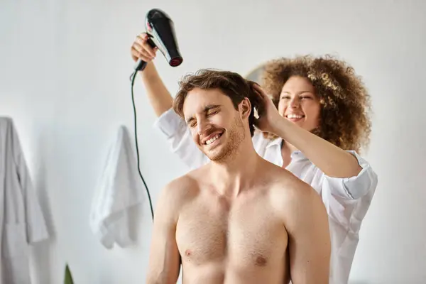 Hombre sonriendo mientras su novia se seca el pelo con secador de pelo en el baño - foto de stock