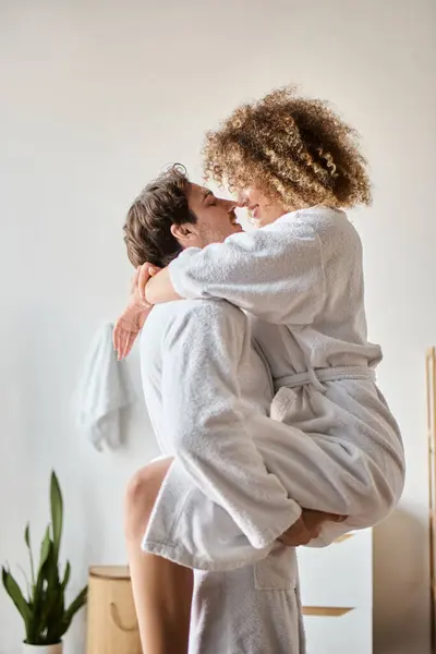 Casal feliz em roupões de banho se divertindo abraçando no banheiro, homem segurando mulher nos braços — Fotografia de Stock