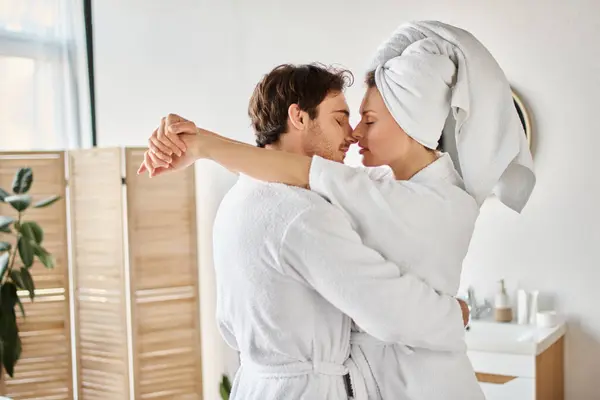 Pareja feliz en albornoces besándose en amor abrazándose en el baño, mujer con toalla en la cabeza - foto de stock