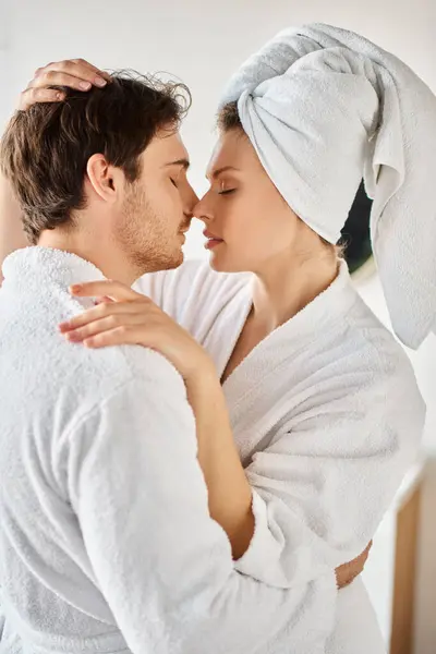 Atractiva pareja en albornoces besándose en amor abrazándose en el baño, mujer con toalla en la cabeza - foto de stock
