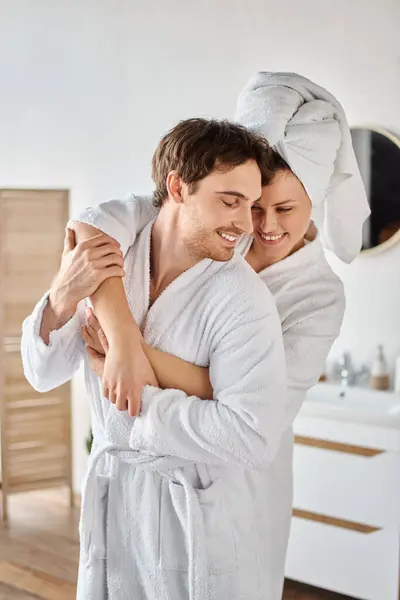 Portrait d'homme souriant en peignoir dans la salle de bain, femme le serrant avec une serviette sur les cheveux — Photo de stock