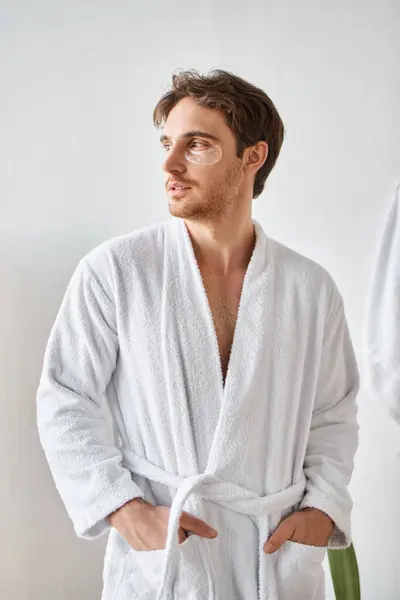 Красивый брюнетка мужчина с повязками на глазу носить белый халат в ванной комнате и смотреть в сторону — стоковое фото