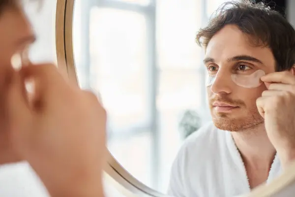 Retrato de hombre moreno con parches en los ojos con bata blanca en el baño y mirándose en el espejo - foto de stock