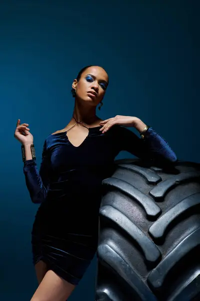 A young African American woman confidently stands next to a massive tire in a studio setting. — Stock Photo