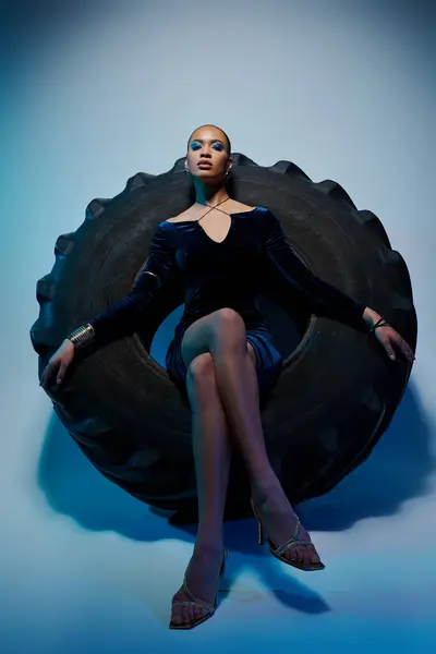 Una joven afroamericana con un vestido negro se sienta en un neumático gigante. - foto de stock