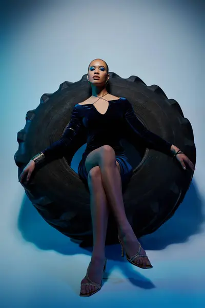 A young African American woman in a black dress sits on a giant tire. — Stock Photo