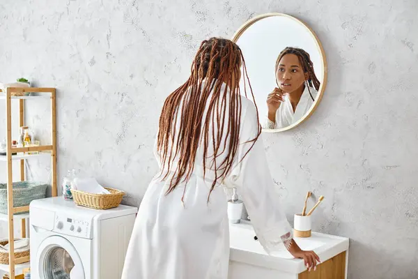 Афроамериканка в халате и афрокосичках чистит зубы перед зеркалом в современной ванной. — стоковое фото