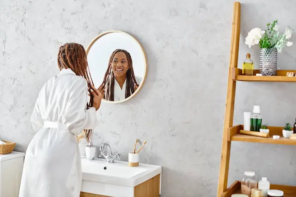 Eine afroamerikanische Frau mit Afro-Zöpfen steht in ihrem modernen Badezimmer und bewundert ihr Spiegelbild im Spiegel. — Stockfoto