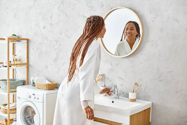 Mulher afro-americana em roupão de banho com tranças afro fica na frente da máquina de lavar roupa moderna no elegante banheiro. — Fotografia de Stock