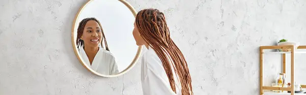 An African American woman with afro braids gazes at her reflection in a mirror in a modern bathroom, contemplating beauty and hygiene. — Stock Photo