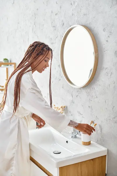 Una mujer afroamericana con trenzas afro se lava las manos en un baño moderno, practicando la higiene personal y el autocuidado. - foto de stock