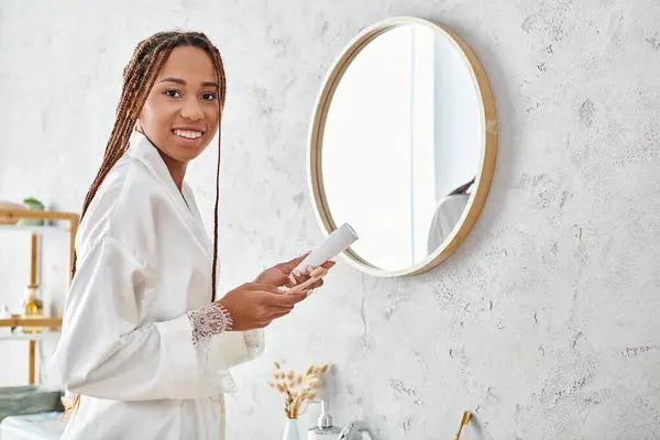 An African American woman with afro braids in a bathrobe is diligently brushing in her modern bathroom. — Stock Photo