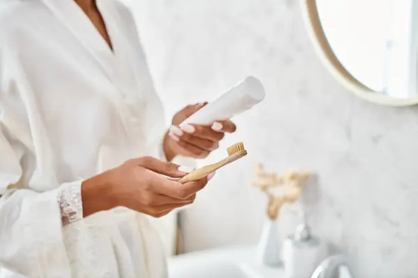 Uma mulher afro-americana em um manto branco tem ternamente uma escova de dentes de cabelo, cercada por um ambiente de banheiro moderno. — Fotografia de Stock