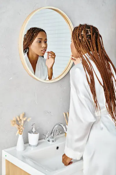 Eine afroamerikanische Frau mit Afro-Zöpfen putzt sich in einem modernen Badezimmerspiegel die Zähne, während sie einen Bademantel trägt. — Stockfoto