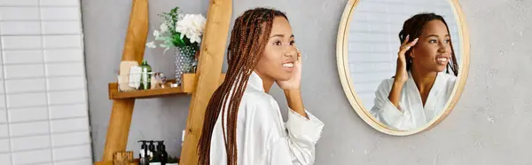 An African American woman with afro braids stands in front of a mirror, brushing her hair in a modern bathroom. — Stock Photo