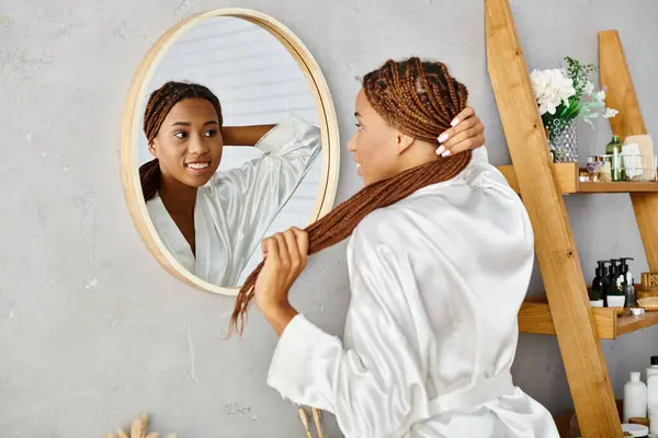 A stylish African American woman with afro braids brushes her hair in front of a mirror in a modern bathroom. — Stock Photo