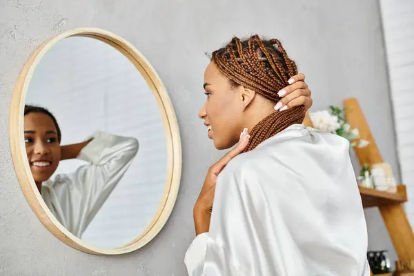 A woman with afro braids stands in front of a mirror in a modern bathroom, brushing her hair in a bath robe. — Stock Photo