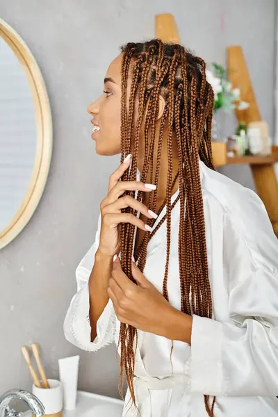 Une Afro-américaine avec des tresses afro se brossant les cheveux dans une salle de bain moderne tout en portant un peignoir de bain. — Photo de stock