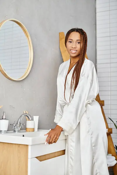 Eine afroamerikanische Frau mit Afro-Zöpfen steht elegant an einem modernen Waschbecken im Badezimmer und konzentriert sich auf Schönheit und Hygiene. — Stockfoto