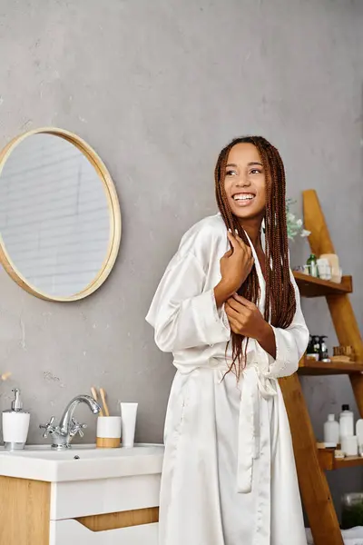 Donna afro-americana con trecce afro in accappatoio, in piedi di fronte al moderno lavabo da bagno, concentrandosi sulla bellezza e l'igiene. — Foto stock