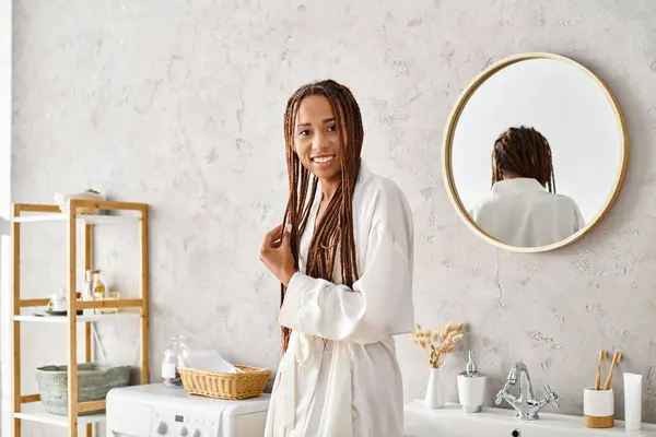 Африканська американка з афро косами стоїть перед дзеркалом у сучасній ванній кімнаті, одягнена в халат. — стокове фото