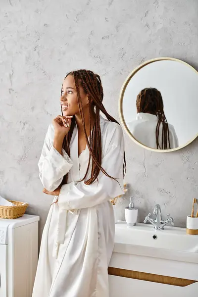 Afroamerikanerin mit Afro-Zöpfen steht im Bademantel vor dem modernen Badezimmerspiegel. — Stockfoto