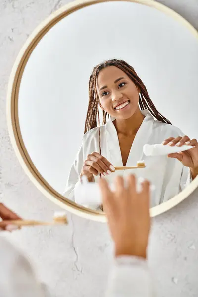 Афроамериканка с афрокосичками в халате чистит зубы перед зеркалом в современной ванной. — стоковое фото