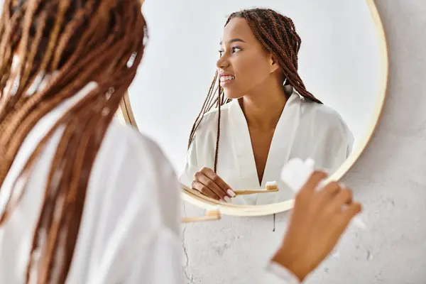 Афроамериканка в халате с афрокосичками чистит зубы перед зеркалом в современной ванной. — стоковое фото