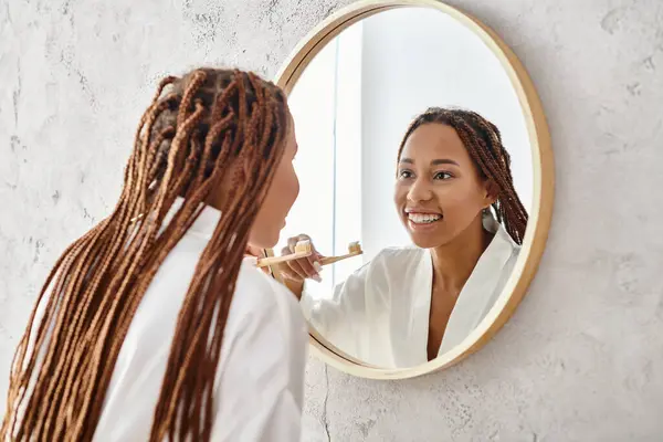 An African American woman with afro braids in a bath robe brushing her teeth in a modern bathroom mirror. — Stock Photo