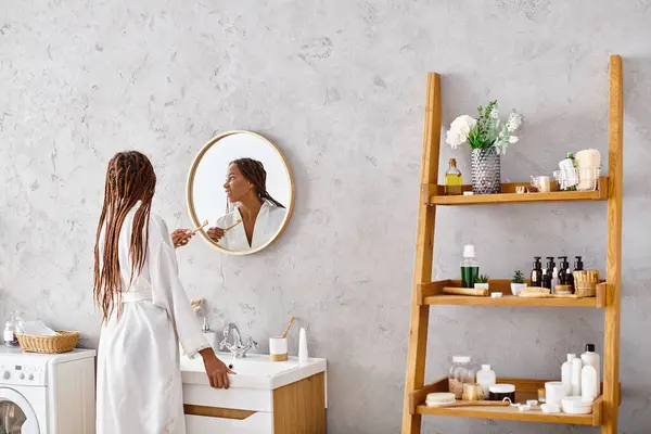 Une femme afro-américaine élégante dans un peignoir avec des tresses afro debout devant un lavabo de salle de bains moderne. — Photo de stock