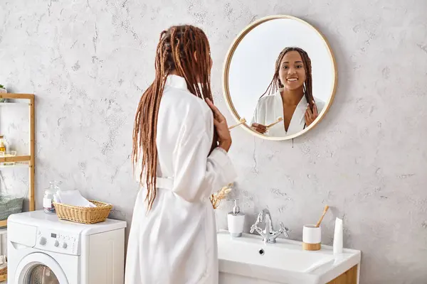 Африканська американка з афро косами стоїть перед дзеркалом у сучасній ванній кімнаті, одягненій у халат. — стокове фото