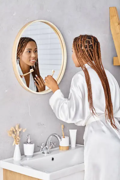 Eine afroamerikanische Frau mit Afro-Zöpfen im Bademantel beim Zähneputzen in einem modernen Badezimmer vor einem Spiegel. — Stockfoto