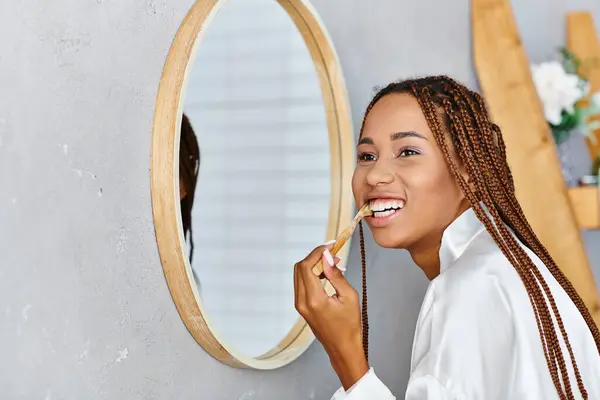 Афроамериканка с афрокосичками в халате чистит зубы перед зеркалом в современной ванной. — стоковое фото