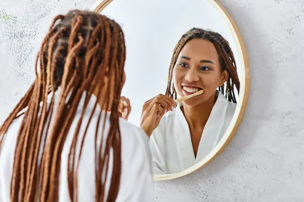 Femme afro-américaine avec des tresses en peignoir de bain brossant les dents devant un miroir dans la salle de bain moderne. — Photo de stock