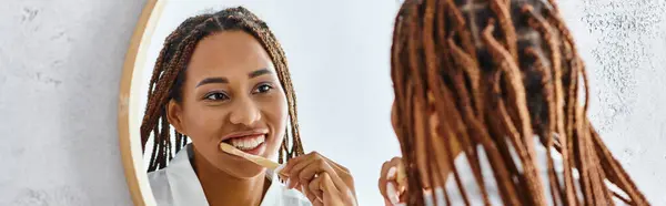An African American woman with afro braids in a bathrobe brushes her teeth in a modern bathroom while looking at her reflection in the mirror. — Stock Photo