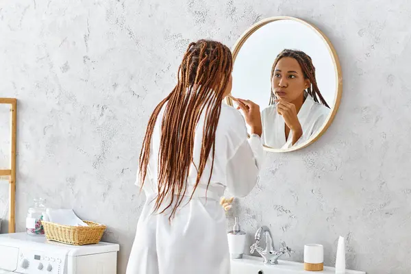 Африканська американка з афро-косами стоїть у своїй сучасній ванній кімнаті, чистить зуби перед дзеркалом. — стокове фото