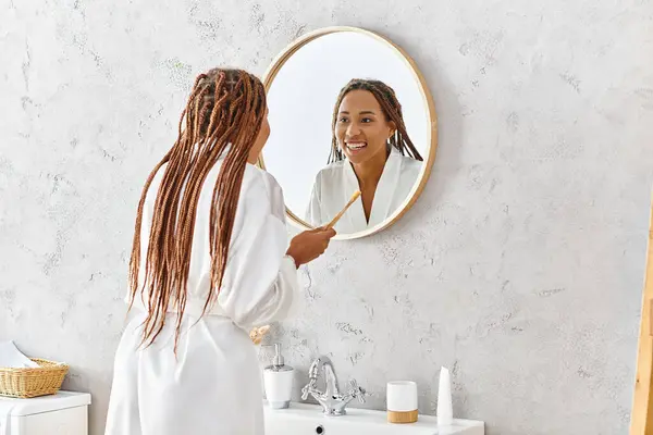 Femme afro-américaine avec tresses en peignoir de bain brossant les dents dans le miroir de salle de bain moderne. — Photo de stock