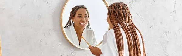 Eine Frau mit Afro-Zöpfen blickt ihr Spiegelbild im Badezimmerspiegel an und konzentriert sich auf Selbstbild und Schönheit. — Stockfoto