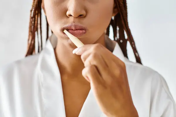 Femme afro-américaine en peignoir avec des tresses afro, se brossant calmement les dents dans la salle de bain moderne. — Photo de stock