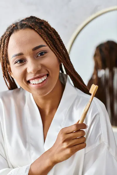 Eine Frau mit Dreadlocks im Bademantel hält eine Zahnbürste in einem modernen Badezimmer und konzentriert sich auf Schönheit und Hygiene. — Stockfoto